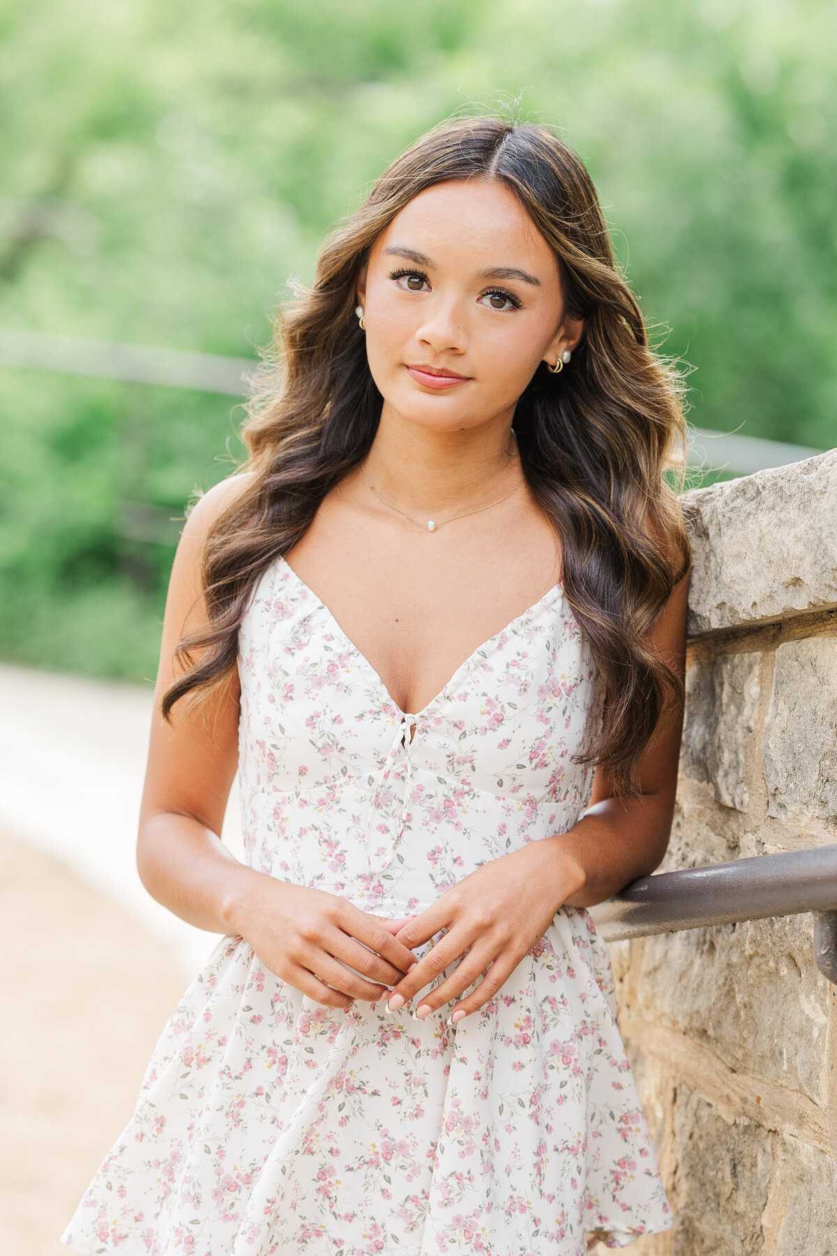 Senior girl with long curled hair leaning on a stone wall, wearing a floral dress and soft makeup, photographed at Arbor Hills in Plano, TX.