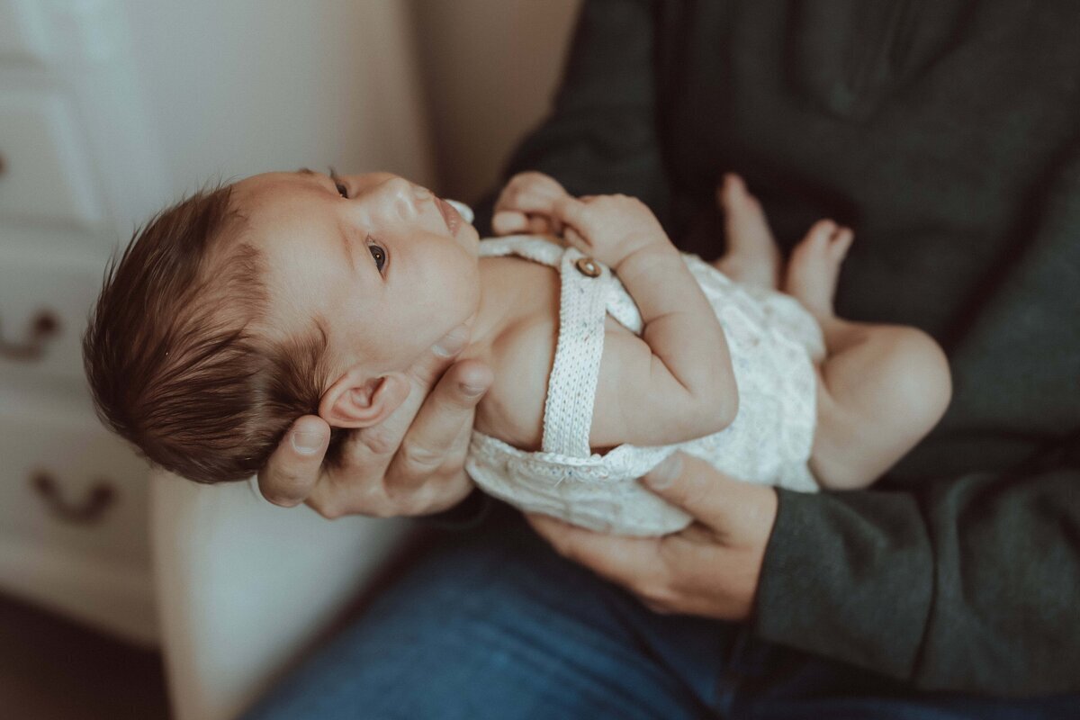 A baby in white overalls is cradled in someone's arms, gazing upward.