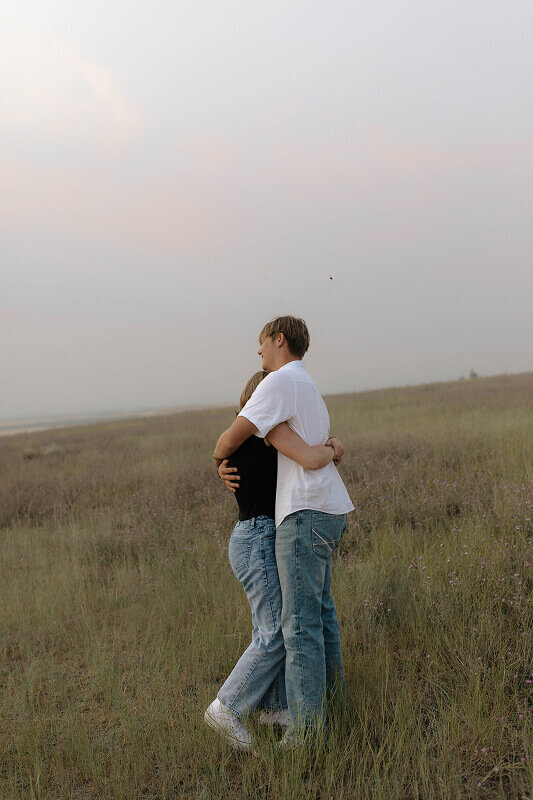 An engaged couple hugs in a  grassy field in Eastern, Idaho. You can't see their faces.