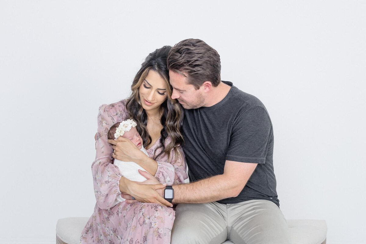A couple sits closely together on a white bench, gazing lovingly at a newborn wrapped in a white blanket. The woman wears a floral dress, and the man wears a black shirt and light pants. The baby has a white headband. The background is plain white.
