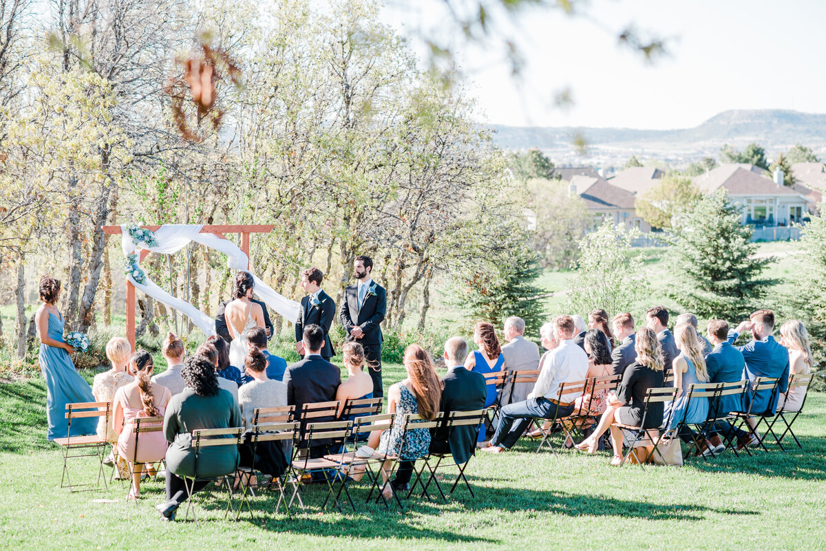 denver wedding photographer captured outdoor wedding ceremony with the guests seated and groom waiting for his bride to arrive as the wind gently blows the trees branches