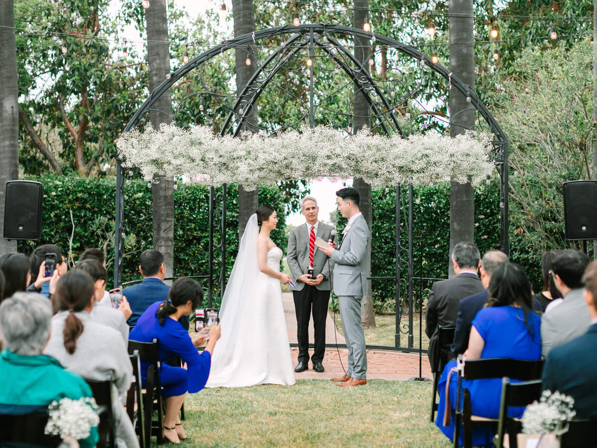 Bride and groom smiling at each other under an arch decorated with white flowers during an outdoor wedding ceremony. Guests are seated and attentive.