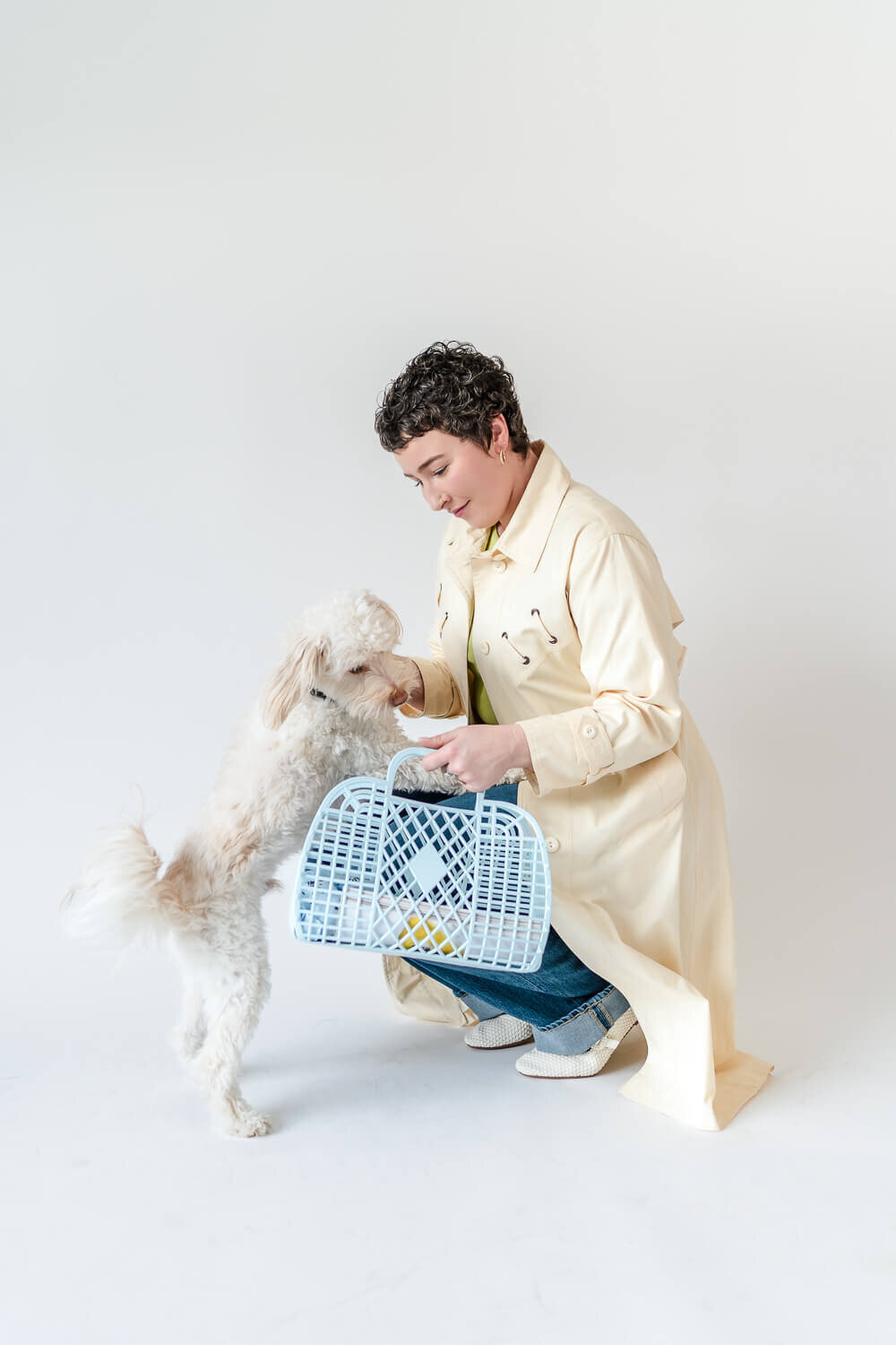 Woman in cream trench coat kneeling to greet white fluffy dog while holding retro blue plastic tote.