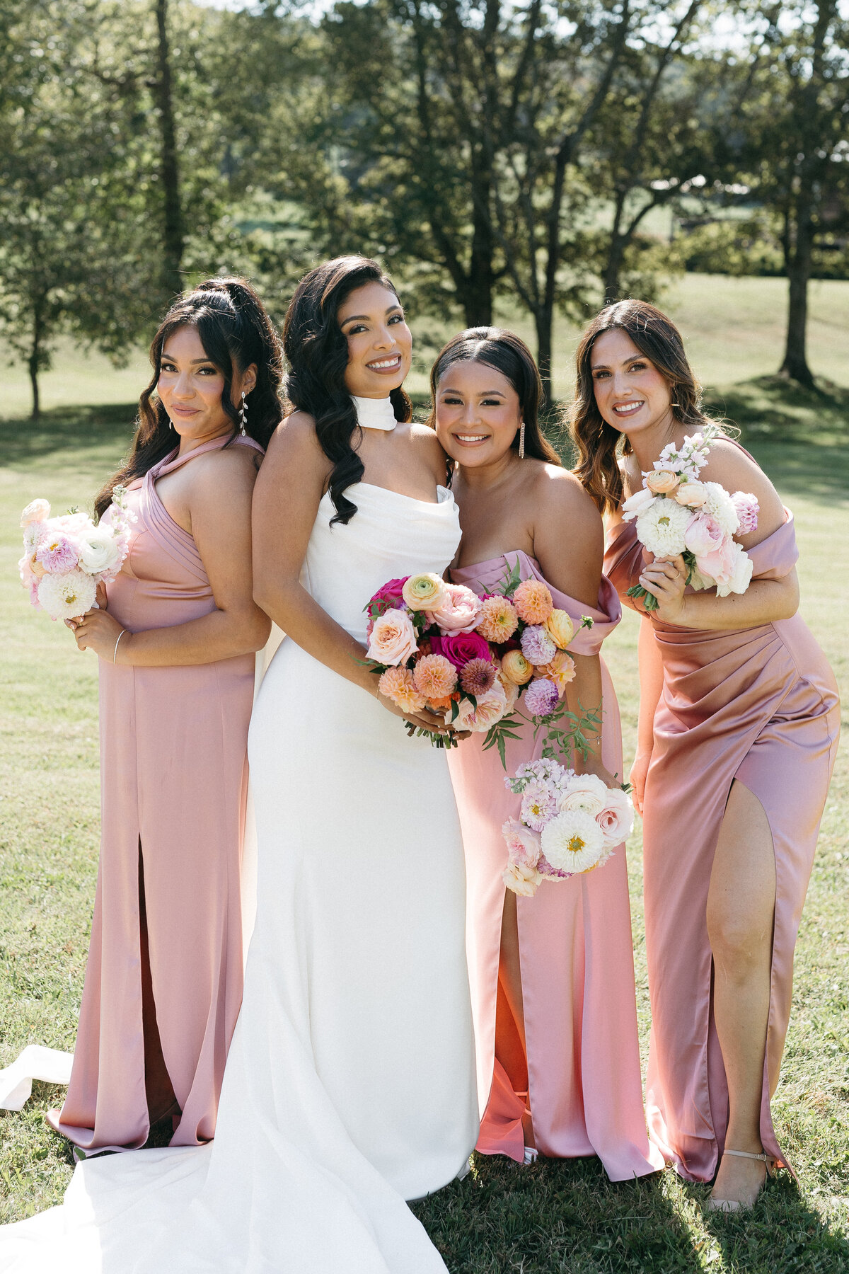 Bride standing with bridesmaids outdoors, all holding pastel garden-style bouquets with roses and dahlias, captured in bright natural light for a soft romantic look.
