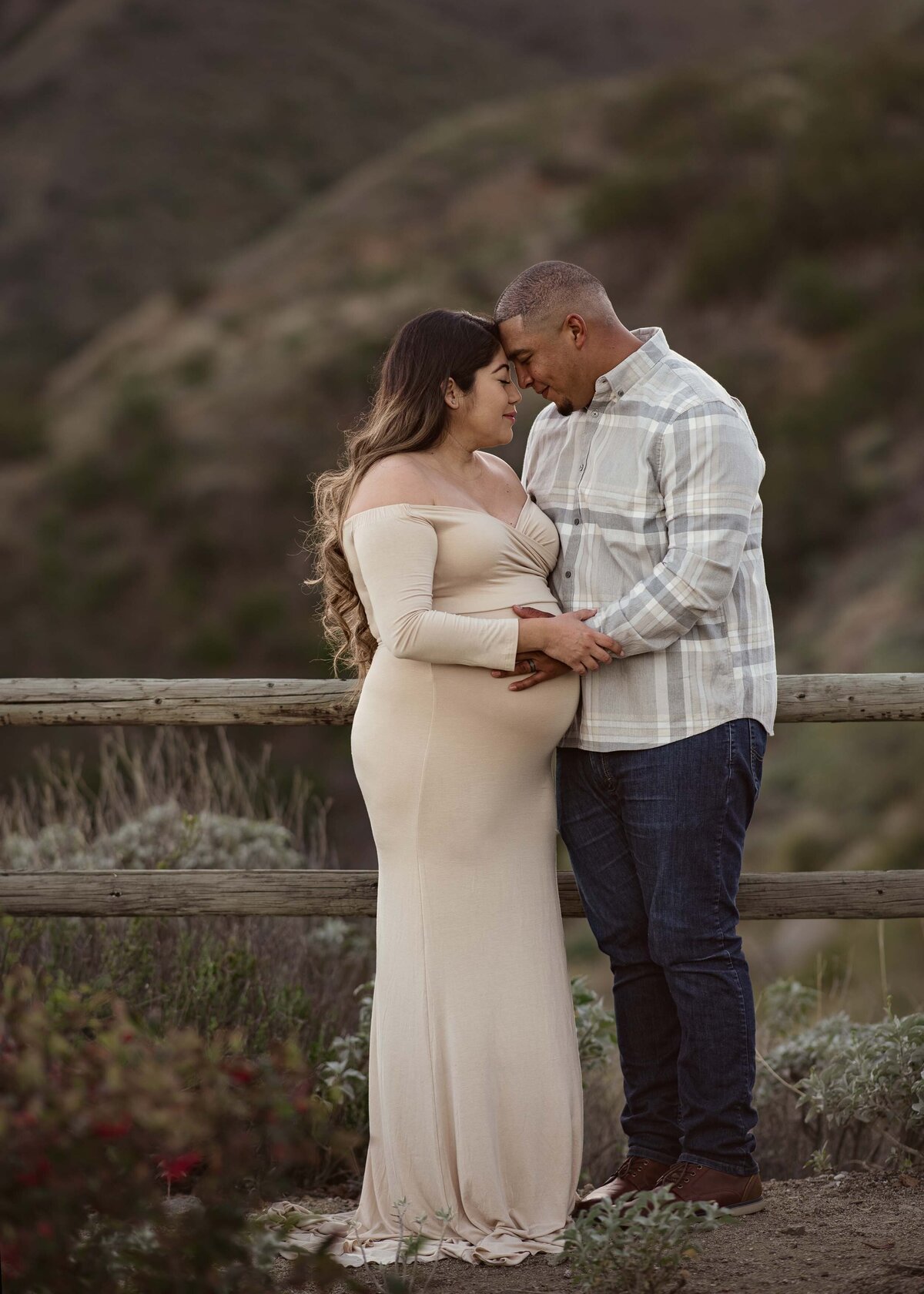 Mom in long, cream, off-the-shoulder maternity gown. Mom and Dad facing each other with their eyes closed and touching their baby bump.
