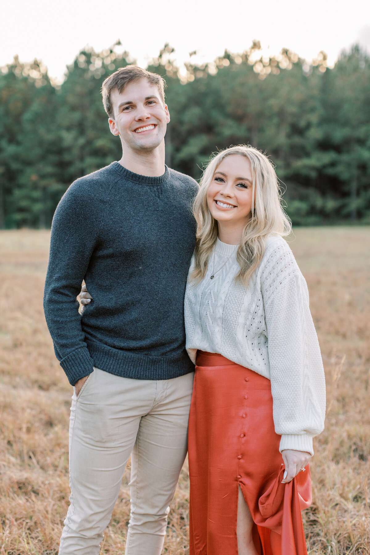 A couple smiles big while standing in a field surrounded by pine trees.