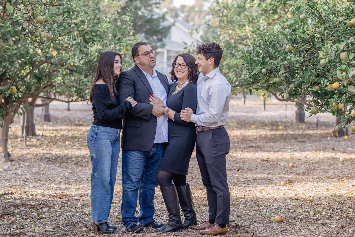 A family of four with dark hair, wearing shades of blue and black standing between a row of green orange trees, laughing and looking at each other. Captured at Irvine Ranch Park.