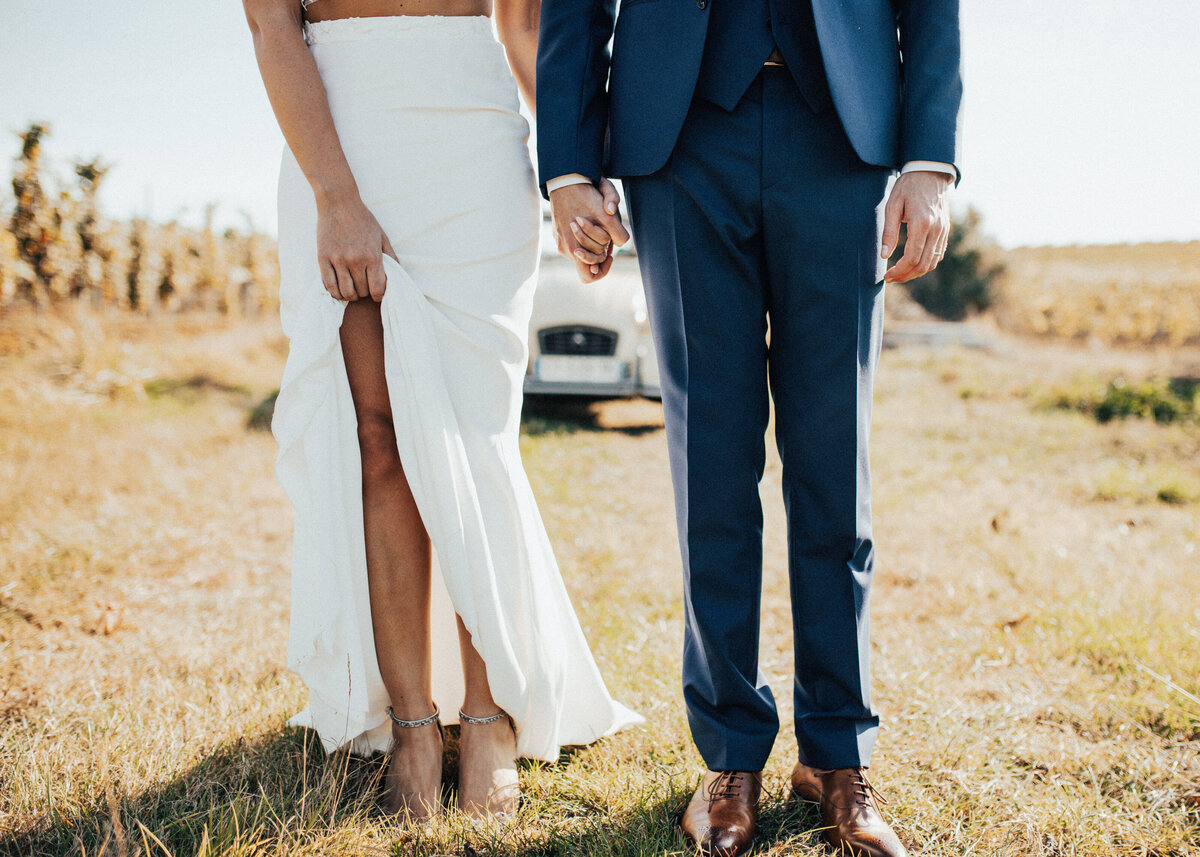 A bride and groom stand in a field with a car behind them. The bride lifts her long bridal skirt.