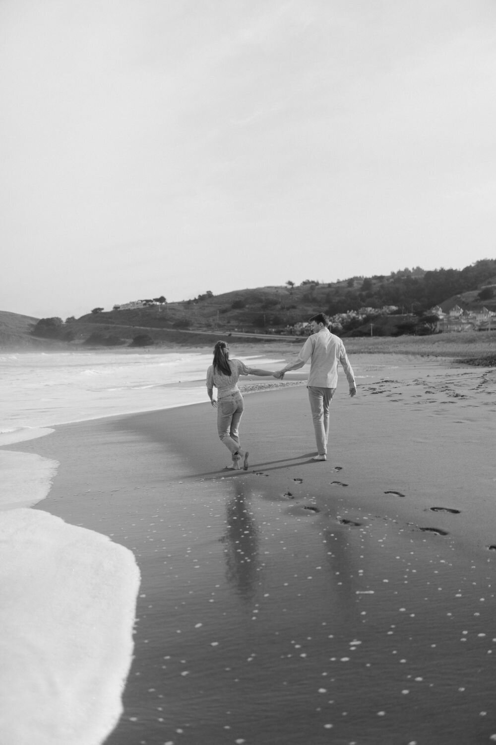 black and white photo of Guy and girl running through beach shore in pacifica california holding hands barefoot