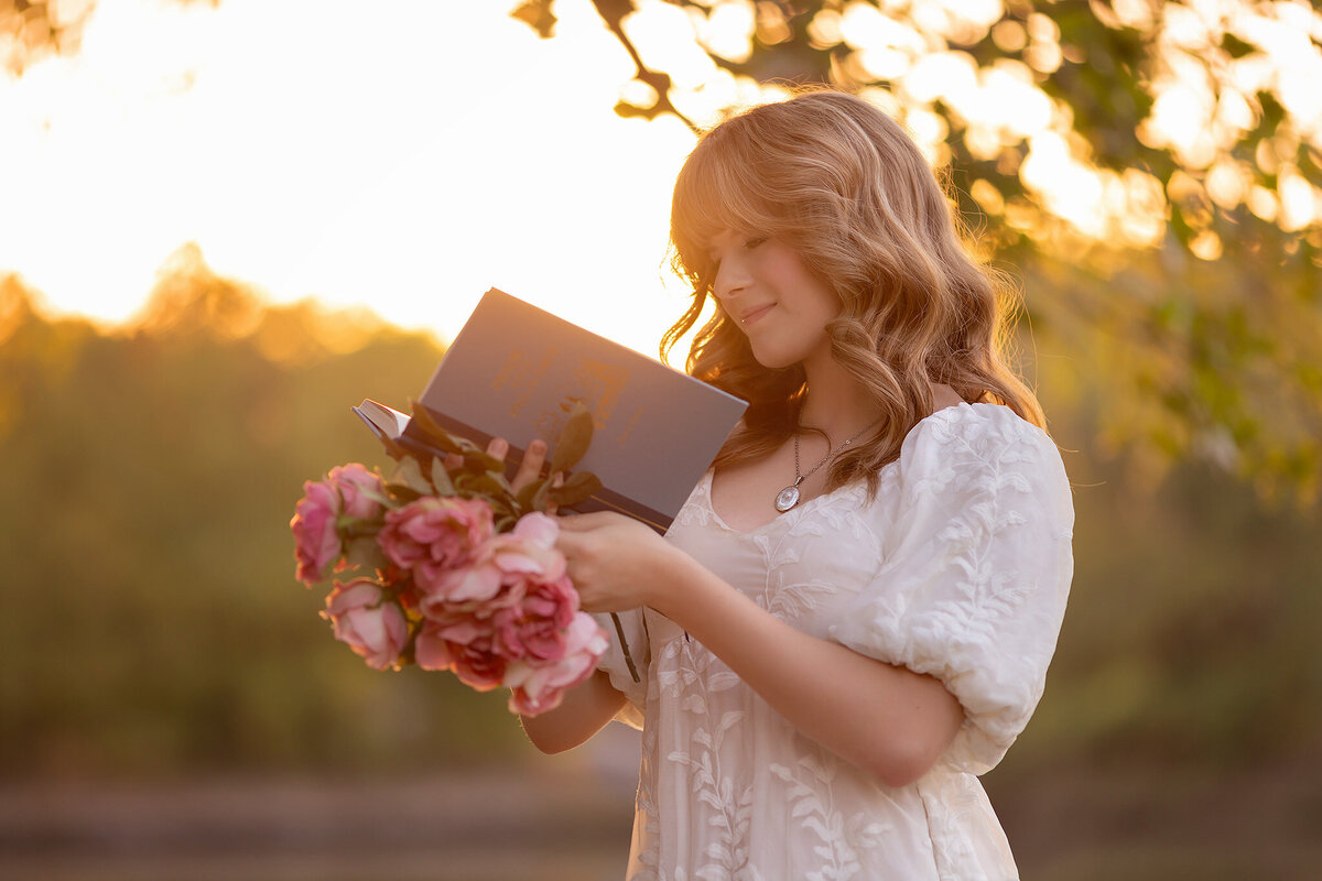 Dallas senior photographer captures a young woman in a floral dress reading a book while holding a bouquet of pink roses, bathed in warm sunset light.