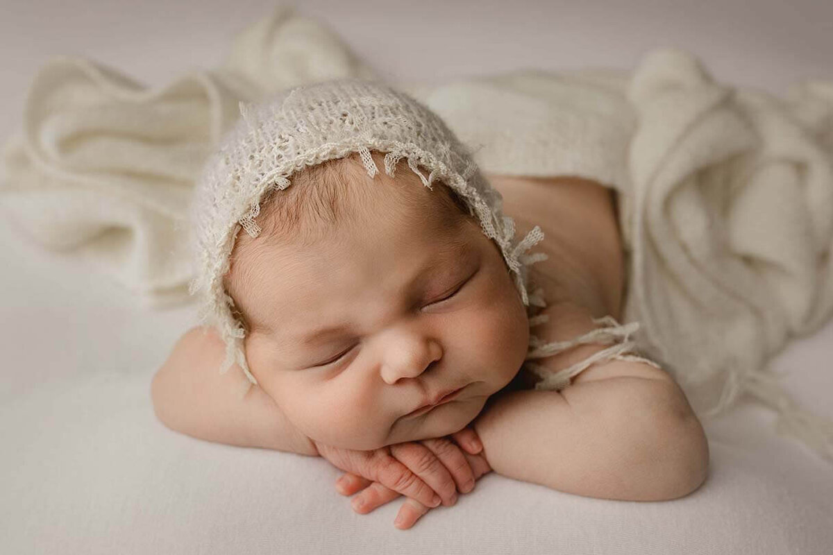 A sleeping baby girl is posed with her hands on her chin while wearing a white bonnet with white fabric draped over her