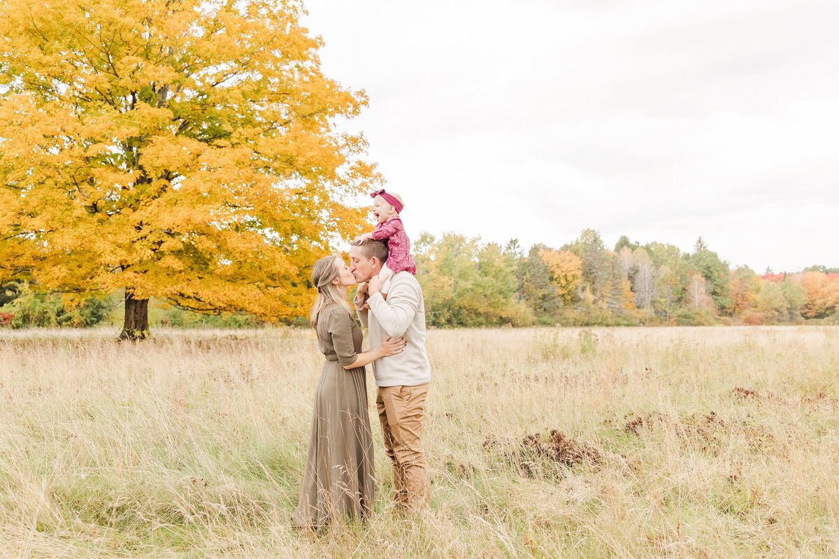 parents kiss in field while daughter sits on dads shoulders during Wayland Massachusetts Family Photo session at Cow Common with Sara Sniderman Photography