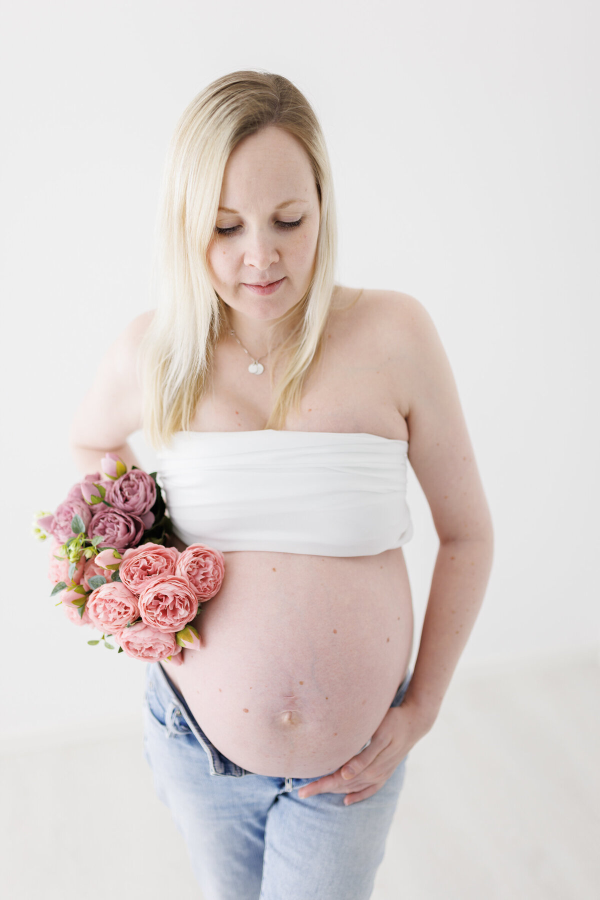 En gravid kvinne fotografert i Studio Landmark. Kvinnen har på blå dongeribukse som er åpen, hvit topp over brystene og en blomsterbukett i høyre hånd. Fotografert i Studio Landmark, Fyllingsdalen.