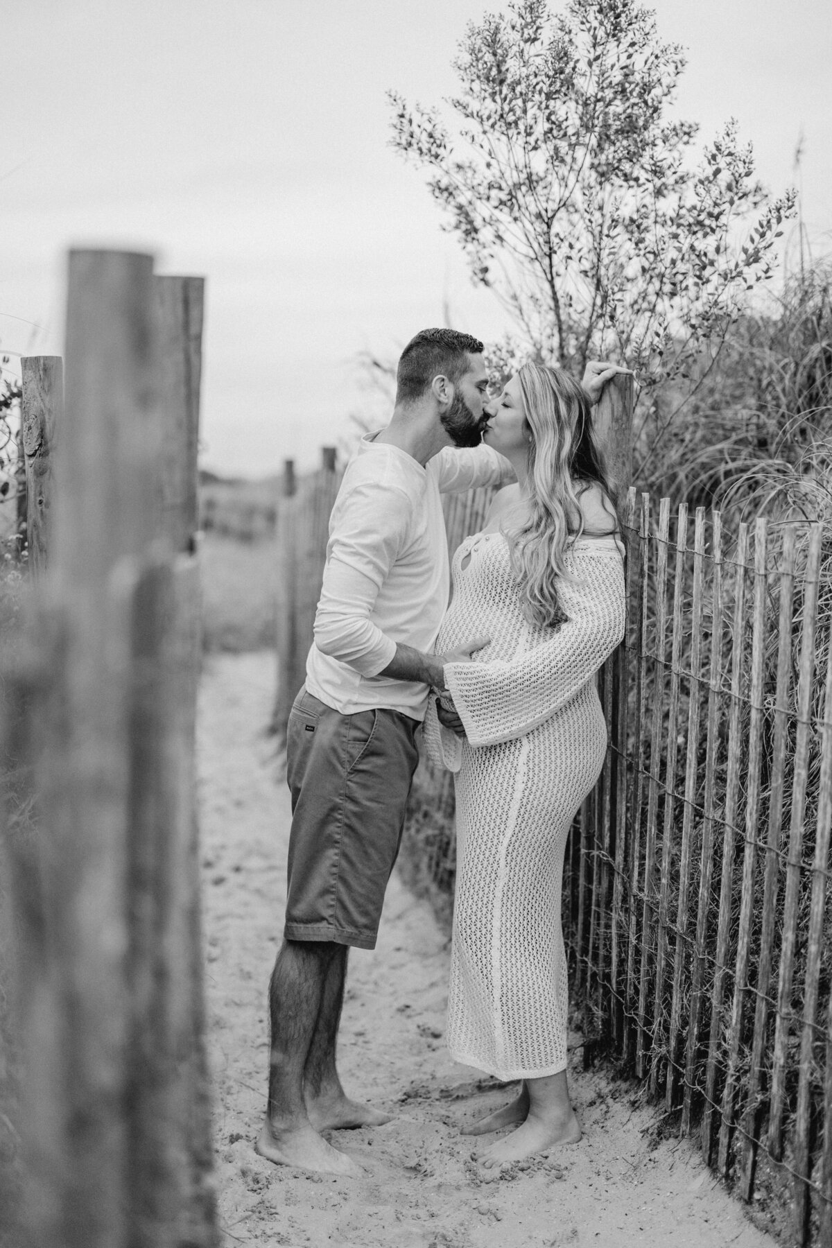 Couple kissing by beach fence in Wilmington, NC, hands lovingly placed on pregnant belly