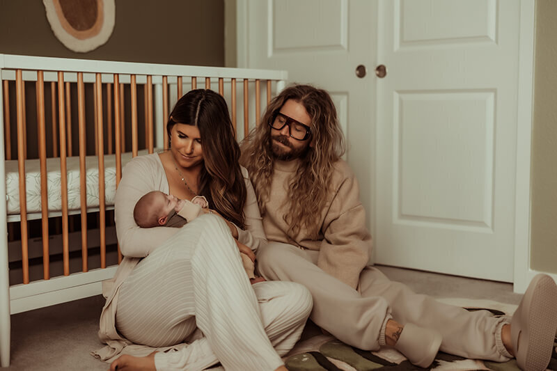 Mom and dad cuddling on the ground in front of crib in nursery while holding newborn baby boy