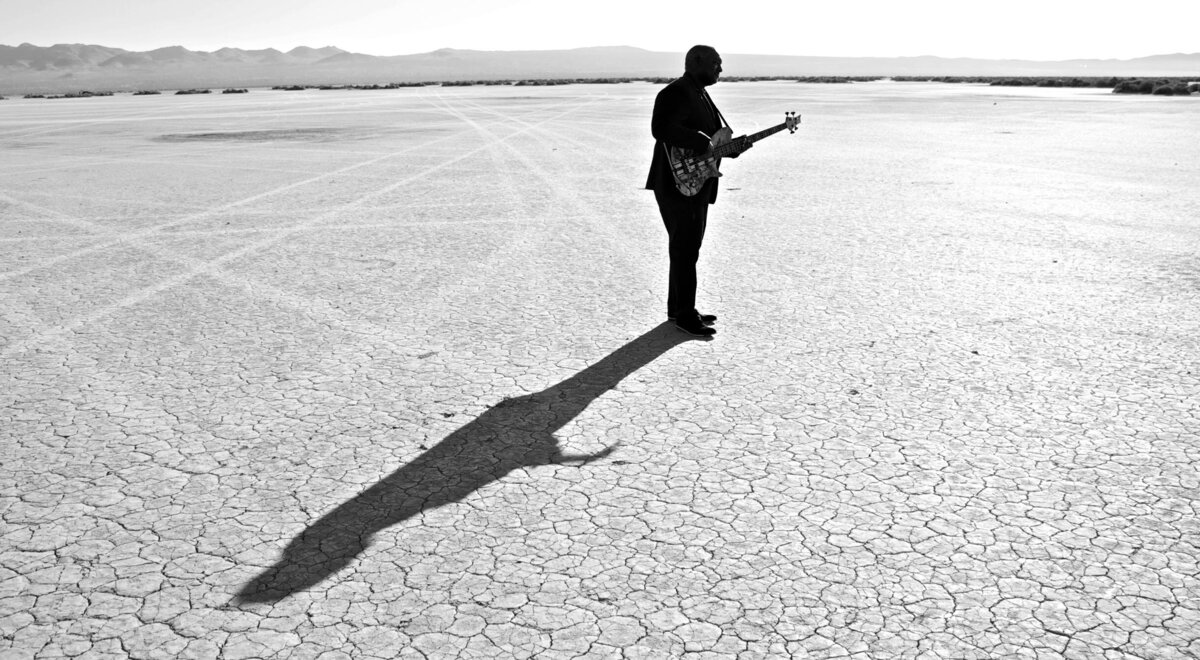 Musician Portrait Mitchell Coleman Jr standing in long shot in desert holding guitar long shadow stretching across sand behind him black and white El Mirage