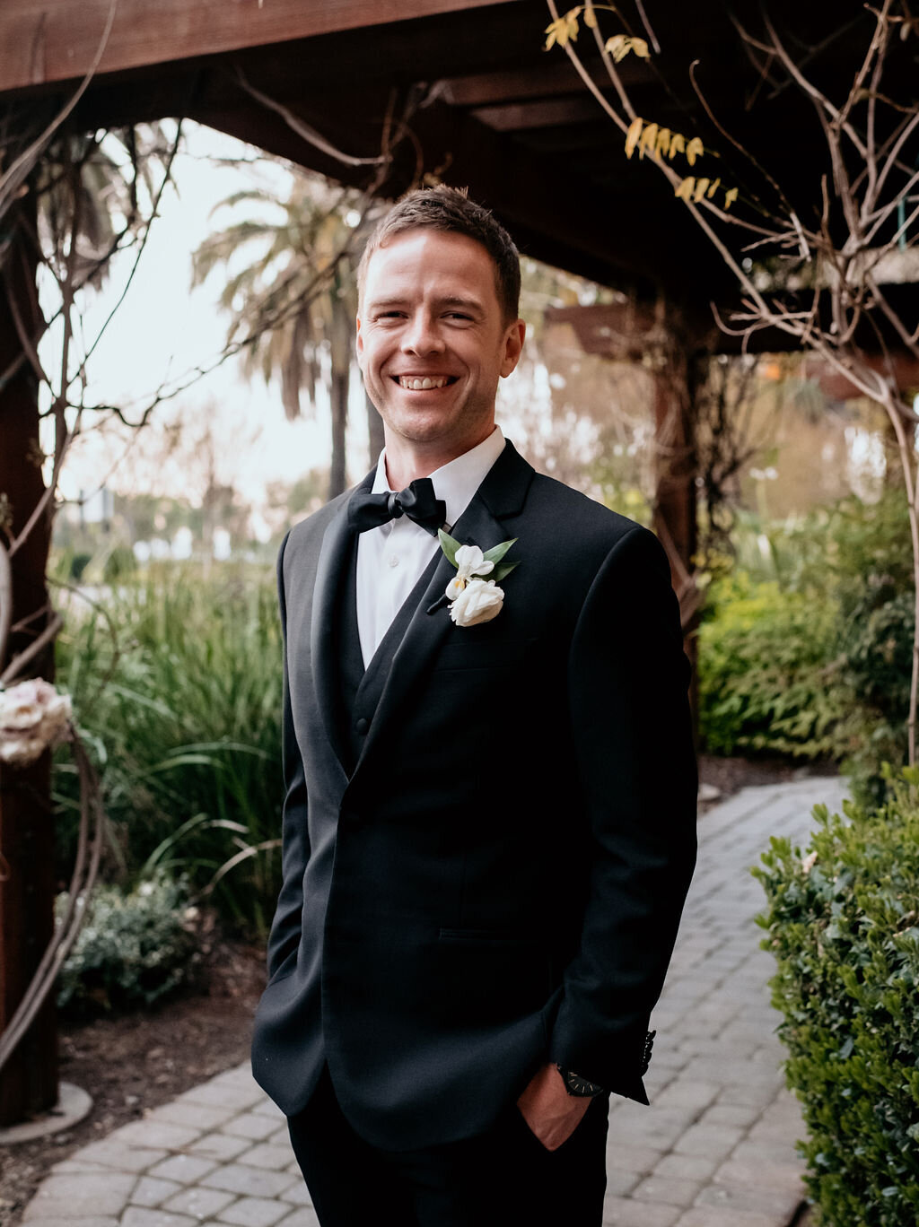 Groom portrait in a classic black tuxedo with a white flower boutonniere, captured at the Palm Event Center in Pleasanton, CA. The spring wedding features a sophisticated color scheme of gold, black, and white, with the groom and his party all dressed in elegant black tuxedos.
