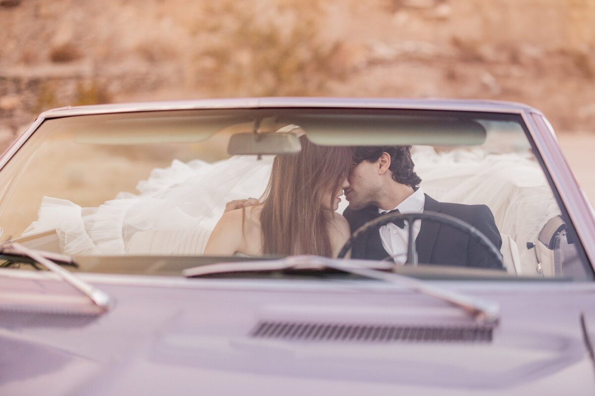 Bride and groom talking in a classic convertible under desert sun.