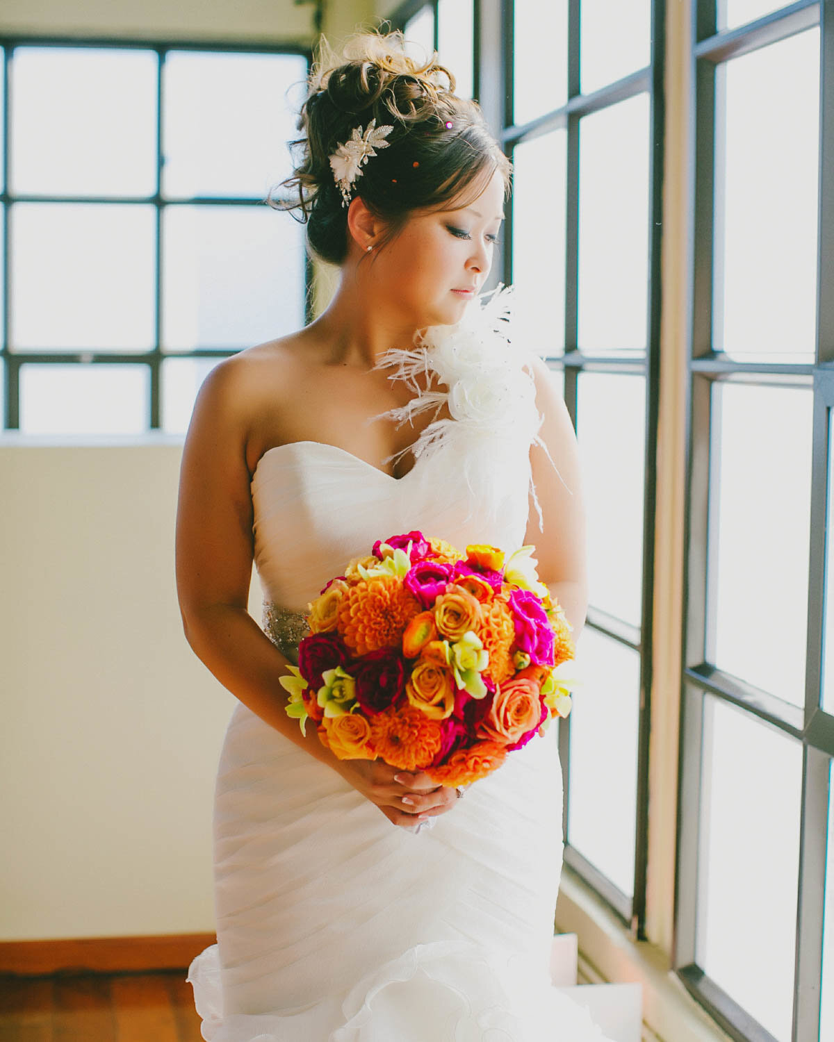Bride holding her bouquet looking down to her right as she stands next to a window