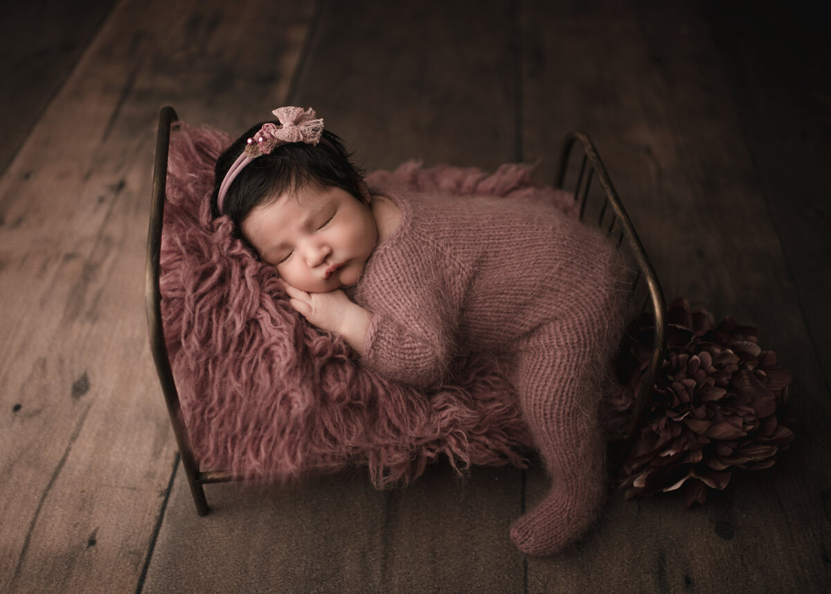 Side profile image of Riverside, CA newborn photoshoot. Baby girl sleeping on her belly on a newborn prop bed with her forearm tucked under her cheek. She is wearing a mauve knit romper. One leg is trailing off the side of the bed. Captured by Best Riverside, CA newborn photographer Bonny Lynn Photography.