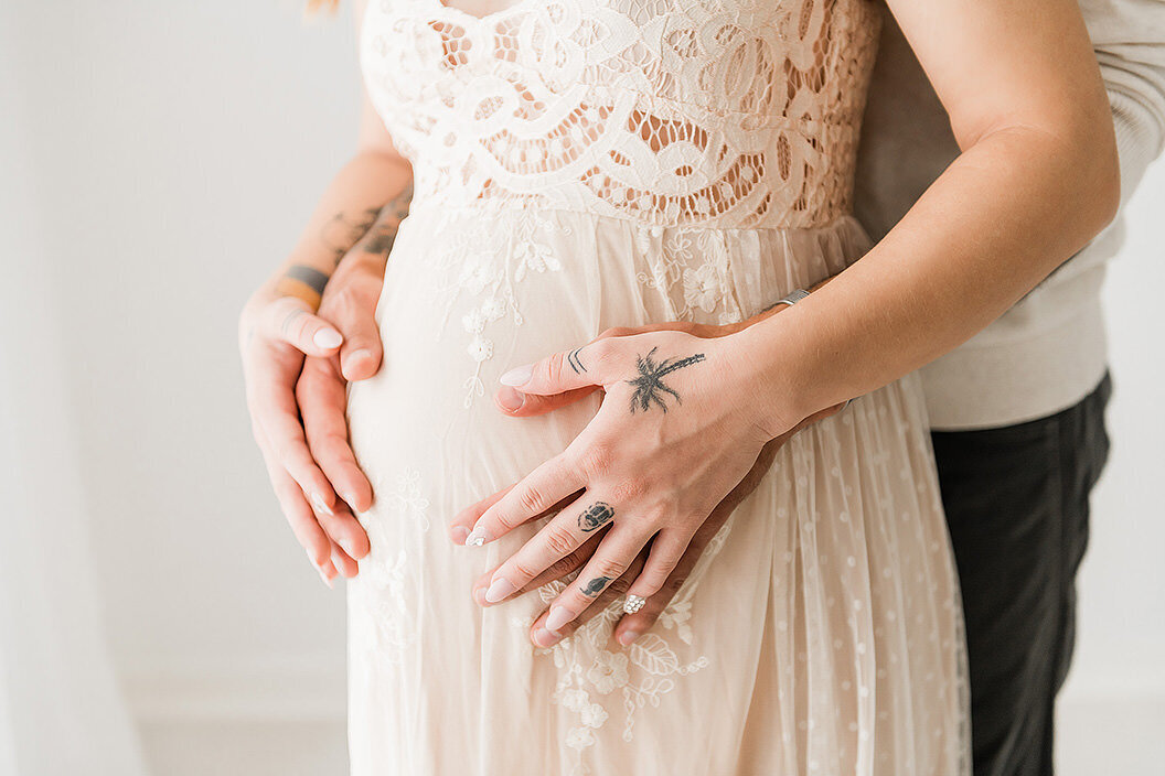 Artistic close-up of tattooed hands resting gently on a pregnant belly in a lace gown, capturing tender connection.