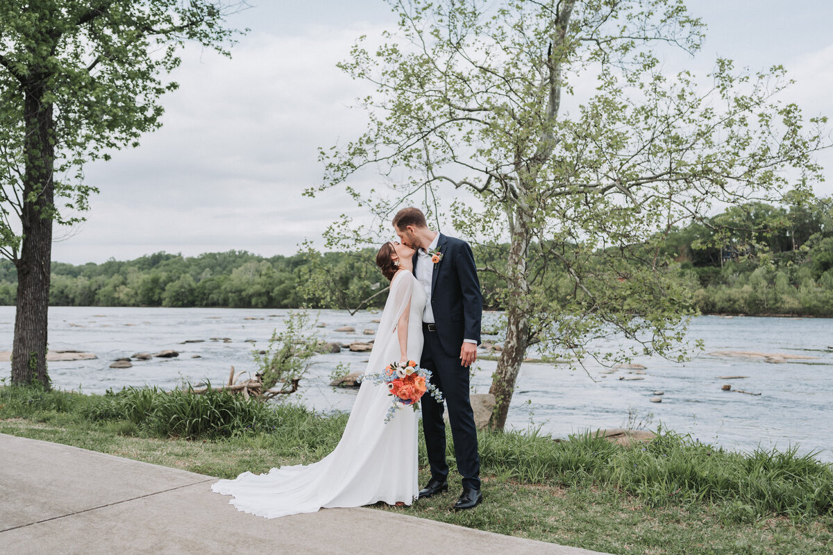 Bride and groom share a kiss beside a scenic river with spring greenery, holding a colorful bouquet in Richmond, Virginia.