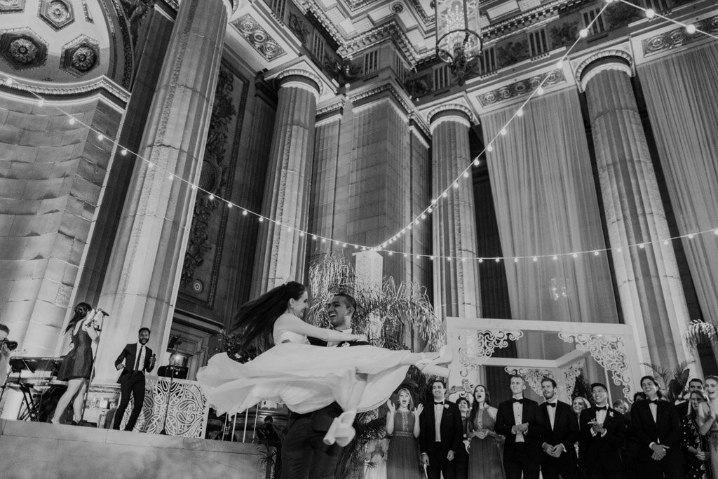 The black-and-white image was captured by photographer Sarah Bradshaw at the Mellon Auditorium in Washington D.C., known as one of the best luxury venues with European flair. The photo depicts a wedding reception in a grand hall with high ceilings and ornate columns. In the center of the picture, the bride and groom are sharing a dance, with the groom lifting the bride into the air, her dress flowing elegantly. Surrounding them are guests dressed in formal attire, watching joyfully in the background.