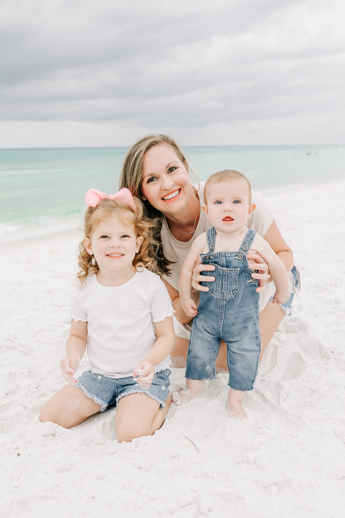 A smiling mother kneels in the soft white sand of Orange Beach, AL, with her two young children. Her curly-haired daughter, wearing a pink bow and denim shorts, plays with the sand while her baby boy, dressed in adorable denim overalls, looks curiously at the camera. The turquoise ocean waves and cloudy sky create a stunning natural backdrop for this joyful beach portrait.