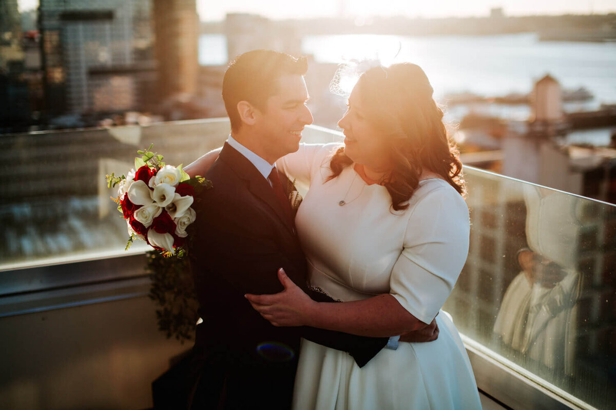 A joyful couple embraces on a rooftop at sunset, with a city skyline and river glowing in the background.