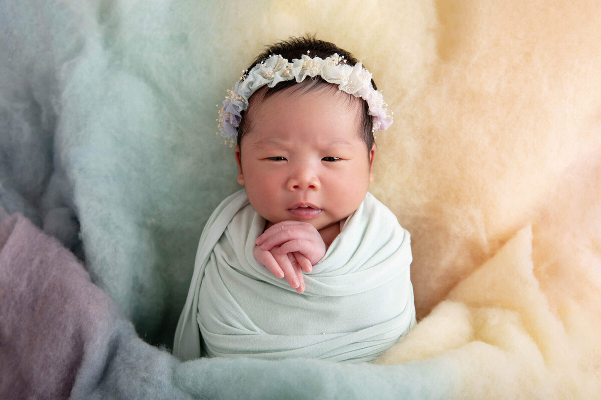 Chinese newborn baby girl wrapped in green laying on a rainbow fluff for her Burlington Newborn Photography Session.