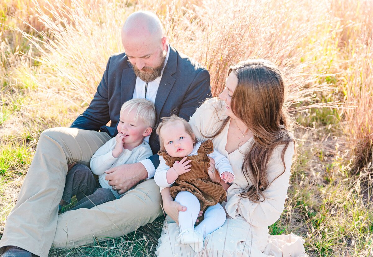 A family with a mom, dad, toddler boy, and infant girl with the kiddos in mom and dad's lap all sit in a field with tall grass and interact with each other captured by denver family photographer