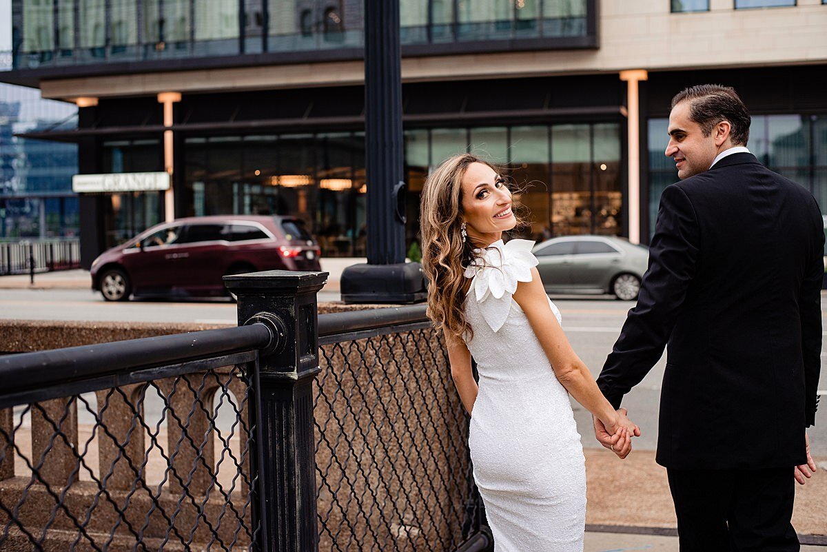 The groom looks at the bride and she looks over her shoulder at the camera. They are walking though  downtown Nashville hand in hand. The bride is wearing a white fitted sheath dress with a ruffle on the shoulder. The groom is wearing a black tuxedo.