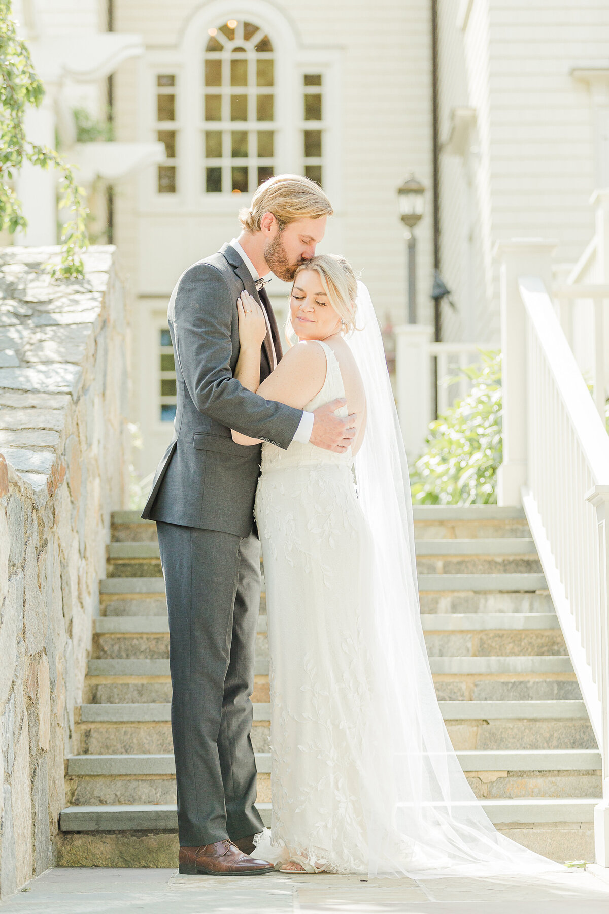 Bride adn groom stand on stone steps in an embrace The bride's hands are on the groom's chest and he his kissing his bride atop of her head. Captured by MA wedding photographer Lia Rose Weddings