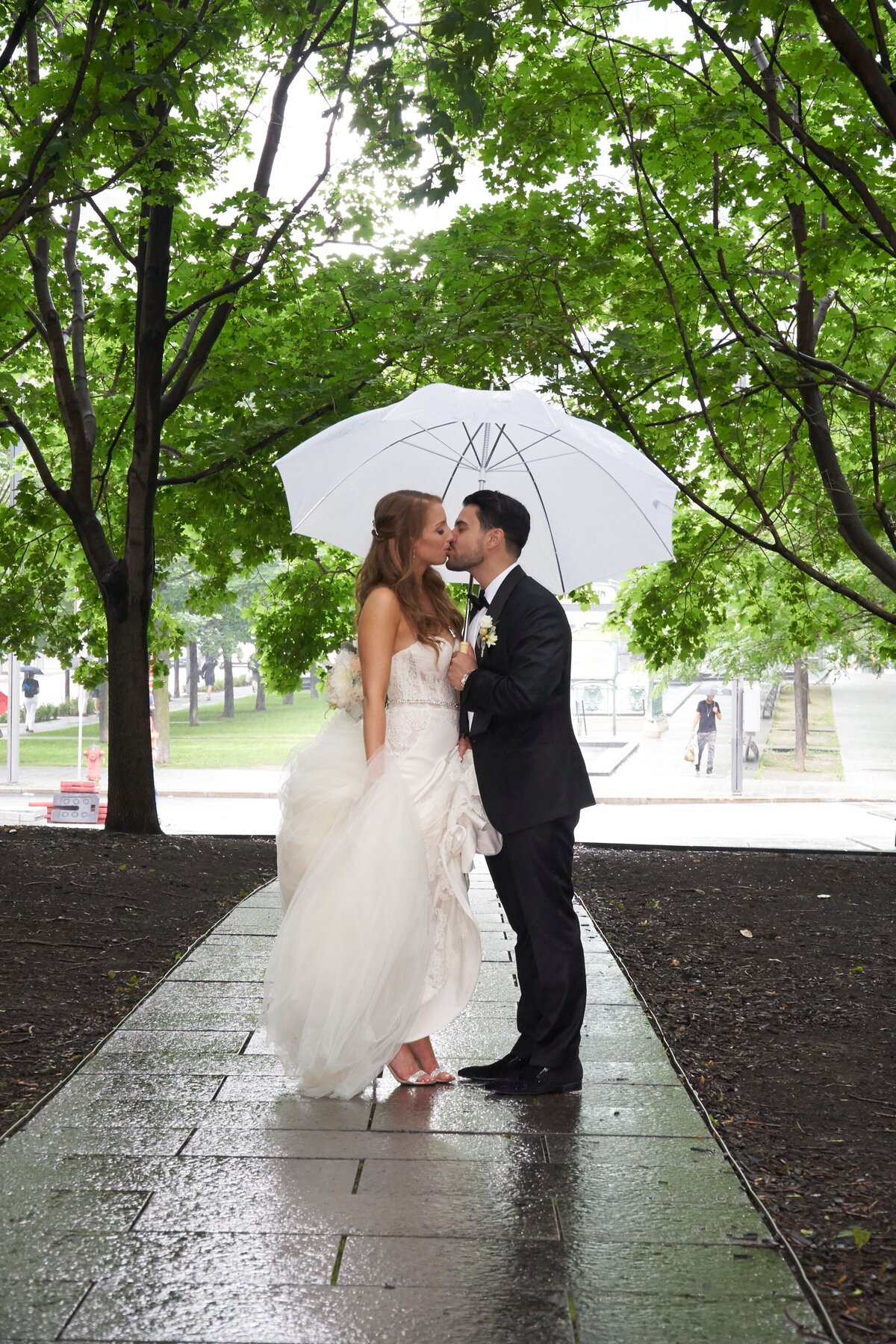 A couple shares a romantic kiss beneath the trees on a beautifully tiled outdoor walkway. The lush greenery and tiled path add a charming and serene backdrop to their affectionate moment.