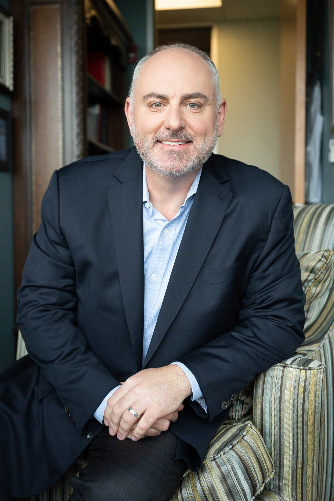 A headshot of a man sitting on a couch with his hands touching and he is smiling to the camera. He is wearing a blue dark suit and a light blue shirt