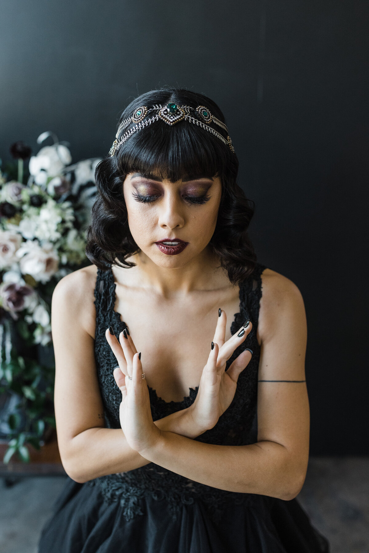 Portrait of a bride on her wedding day in Dallas, Texas. She has her eyes closed and her hands passing by each other as though in deep concentration. She's wearing an intricate black dress,  an ornate headband/tiara, and dark makeup. She poses in front of a bouquet of dark flowers and black background.