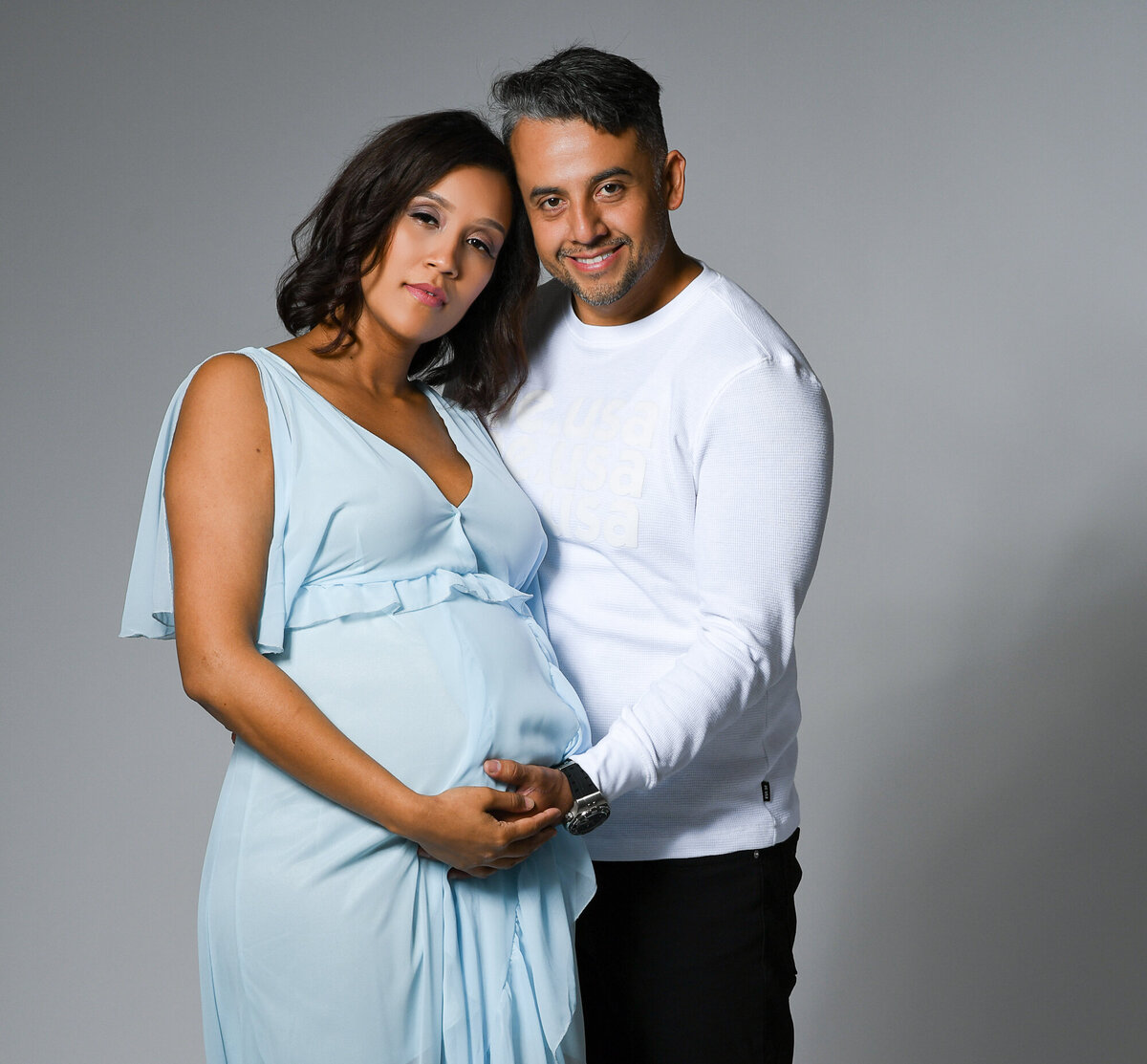Maturnity portrait of woman and her husband holding moms baby bump and smiling into the camera photographed on a grey background