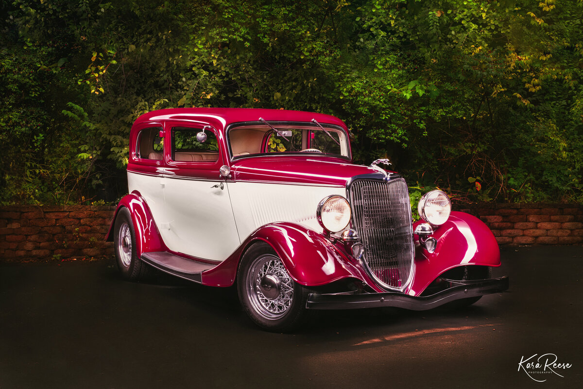 A cherry red and cream 1934 Ford is light painted against a brick and foliage background.