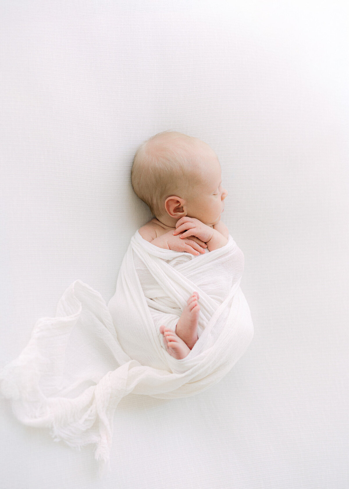Newborn baby boy swaddled in white with his arms and feet out and his head turned to the left while he sleeps durning a session with Indianapolis newborn photographer, Brittney Lear.