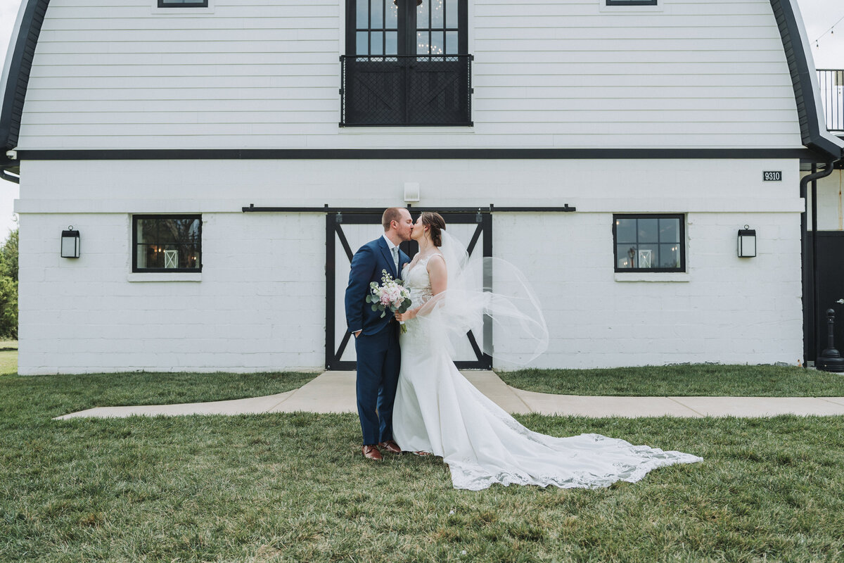 Bride and groom share a kiss in front of a white barn at their Sweeney Barn Virginia wedding, with the bride’s veil floating in the wind.
