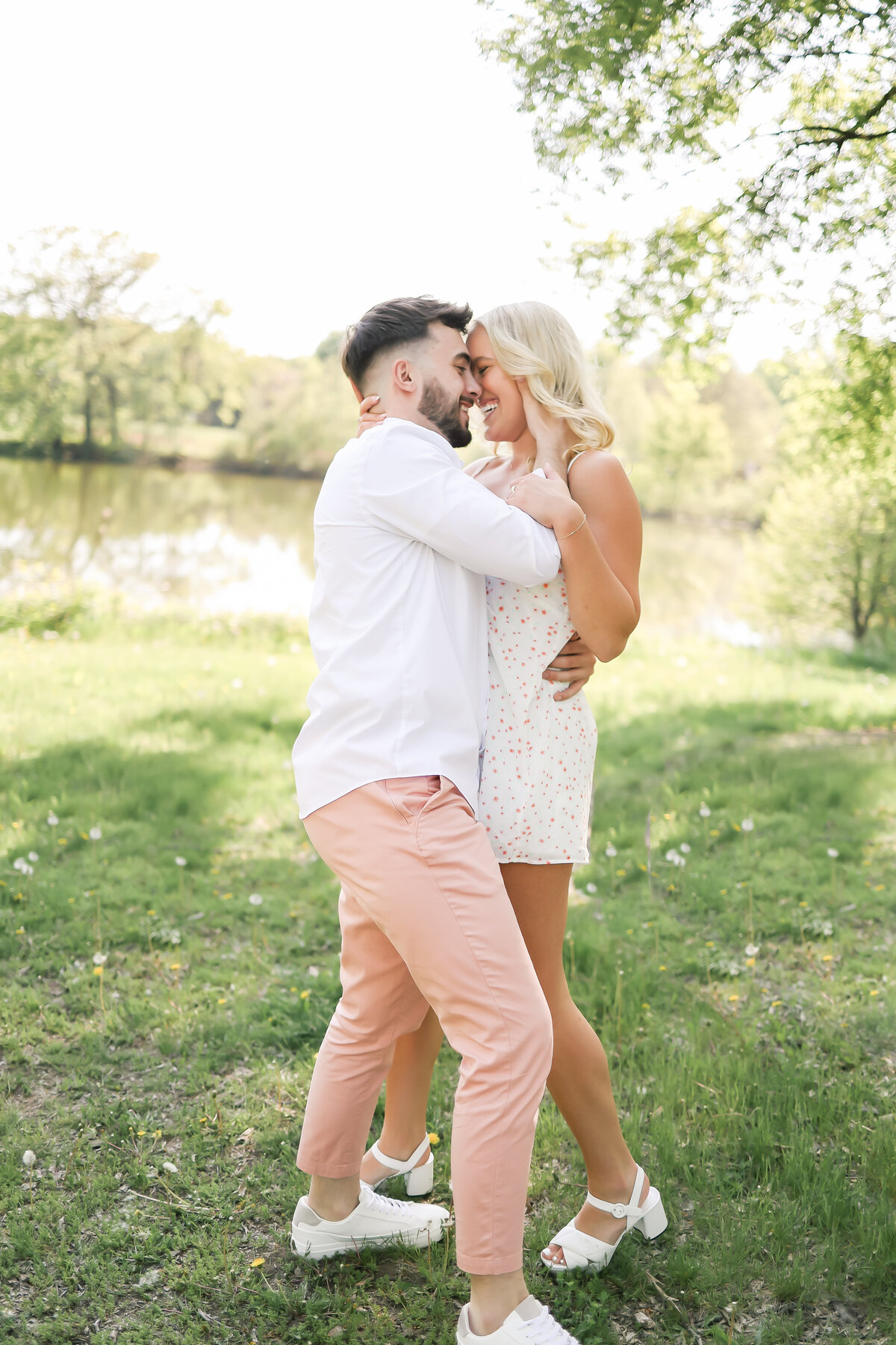 a beautiful couple embrace eachother with a kiss while standing in a field of bright green grass with a lake behind them. the  couple smiles at ecahother as they kiss in Minneapolis, MN