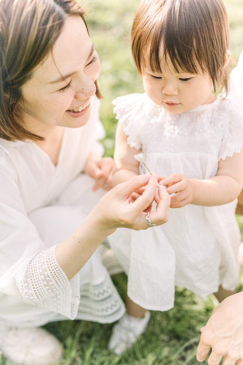 Mother and daughter wearing white dresses are holding a white clover together. Beautiful family photograph shot by Yukika Ishikawa Photography in Shibuya.