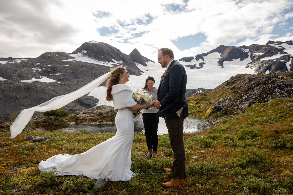 A bride and groom stand facing each other and holding hands during their elopement ceremony in Alaska.