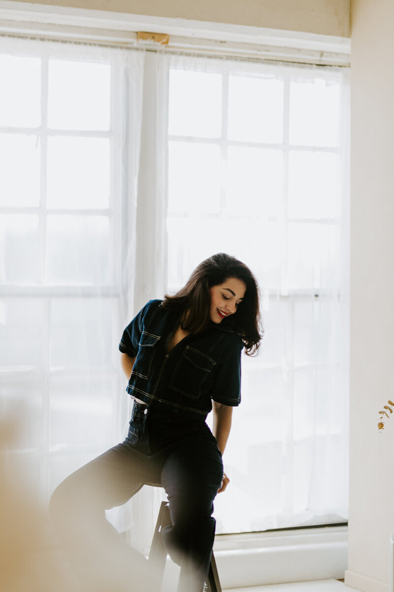 Femme brune, tenue en jean, assise sur un tabouret devant une immense fenêtre lumineuse, posant lors d'une séance photo.