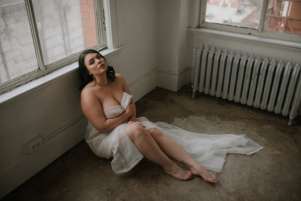 A nude woman holding a white sheet around her poses sitting in front of a window in the Vancouver, BC boudoir studio.