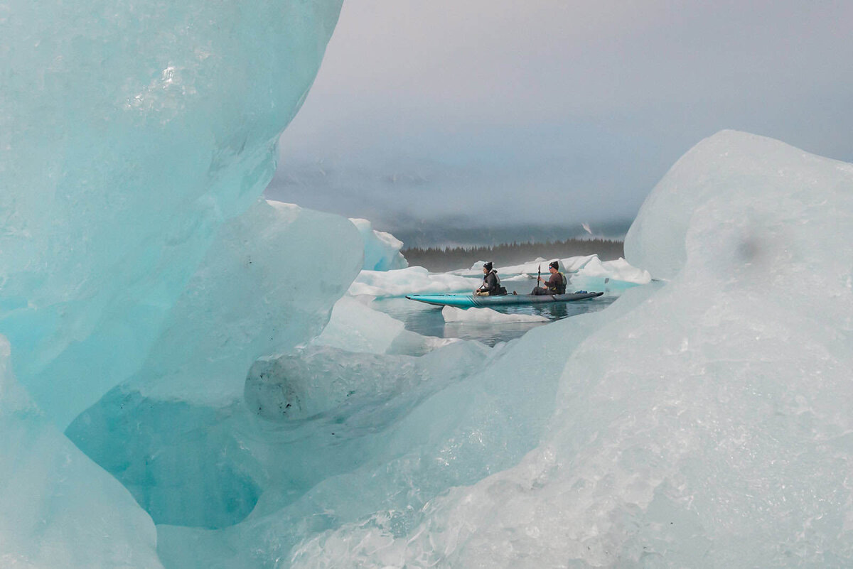 A couple paddles a two person kayak between icebergs curing day two of their Alaska Glacier Elopement.  A large blue and white iceberg naturally framed them in the image as they move across the water. 