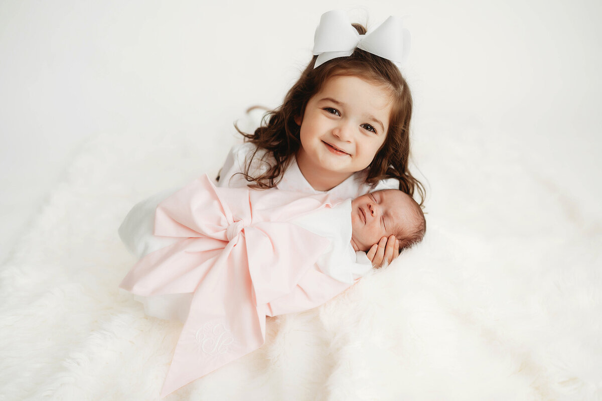Toddler smiles at the camera with her newborn baby sister in her arms during Newborn Photoshoot in Asheville.