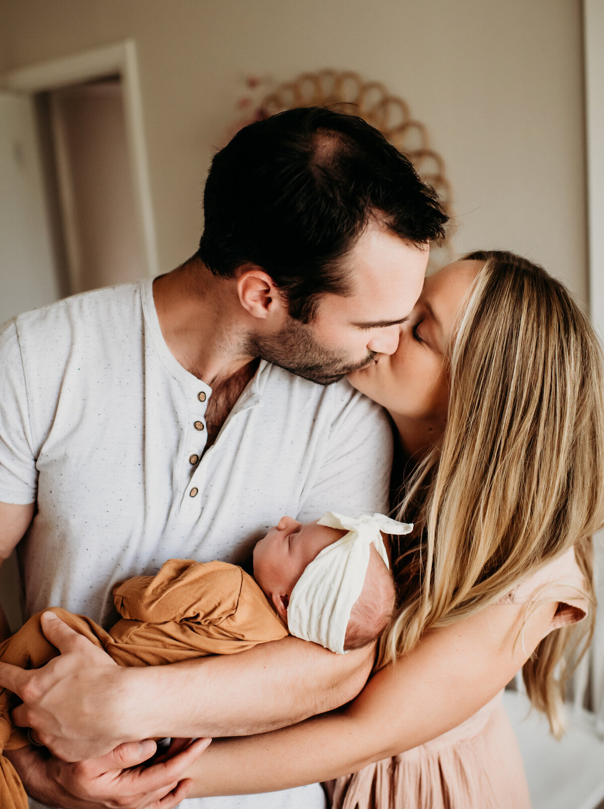Newborn Photographer, a young mother and father kiss in their home as dad holds their new baby