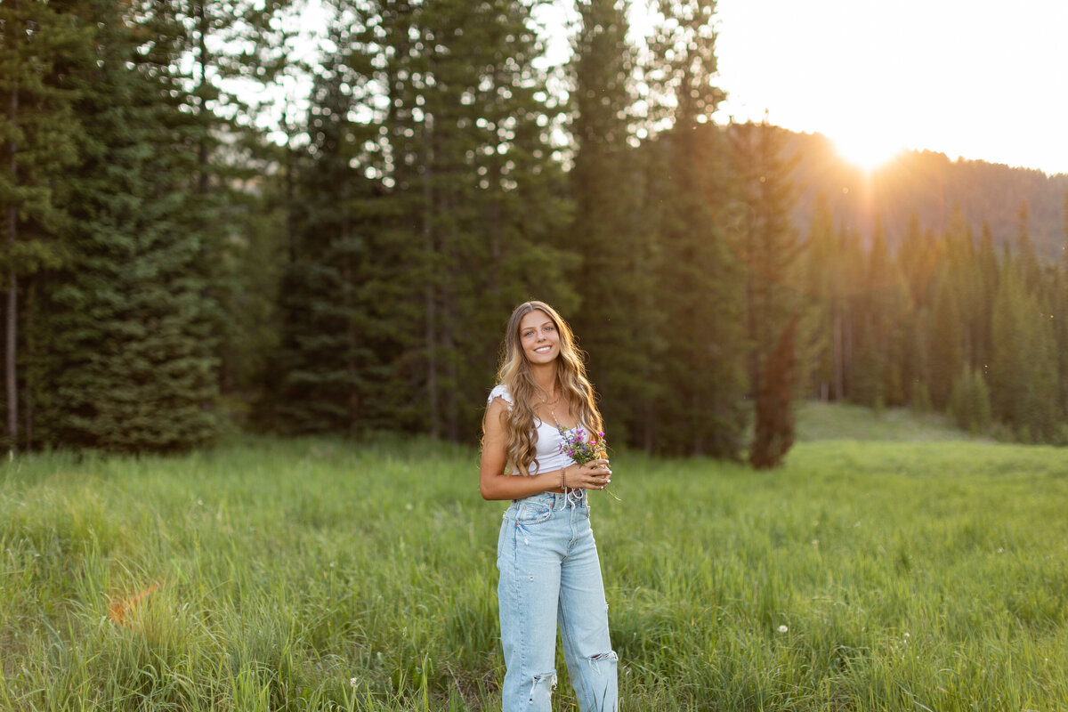 Young woman sits on rocks in a field with large trees as she rests her elbow on her knee.