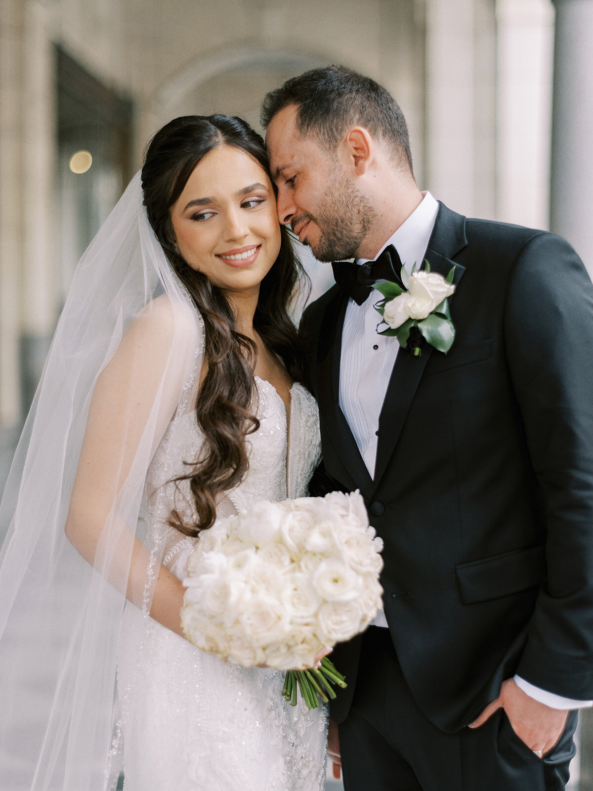 A bride and groom stand close together, the groom leaning his head toward the bride. The bride holds a bouquet of white flowers, wears a white dress and veil, while the groom is in a black tuxedo at a classic Calgary wedding at Fairmont Palliser Calgary.